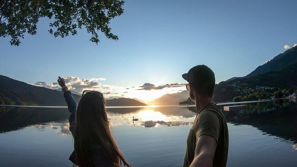 日没時にミルスターター湖の岸に立っているカップル。太陽は高いアルプスの後ろに沈む。湖の穏やかな表面は、オレンジ色の空と山を反映します。彼らは楽しんでいる。日末 - 写真・画像