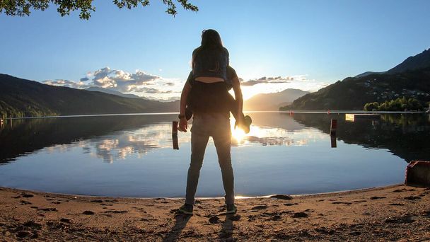 Ein Mann trägt eine Frau huckepack über den Millstätter See während des Sonnenuntergangs. Die Sonne geht hinter den Hochalpen unter. Die ruhige Oberfläche des Sees spiegelt den orangen Himmel wider. Sie haben Spaß, Verspieltheit. Tagesausklang - Foto, Bild