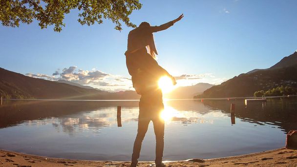 Um homem carregando uma mulher porquinho de volta de Millstaetter lago durante o pôr do sol. O sol põe-se atrás dos Alpes altos. Superfície calma do lago reflete o céu laranja. Eles estão se divertindo, brincando. Fim do dia - Foto, Imagem
