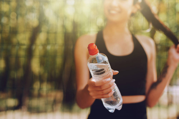 Young woman in black sportswear stretching and doing sport in park. Cropped image of attractive fit girl drinking water on a break. Healthy lifestyle concept. - Foto, Imagen