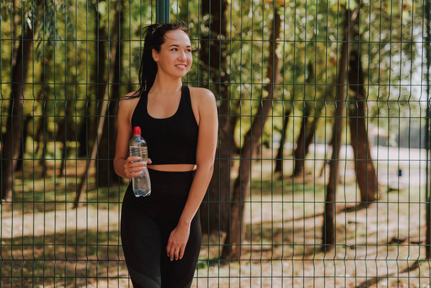 Молодая женщина в черной спортивной одежде растягивается и занимается спортом в парке. Привлекательная приличная девушка пьет воду в перерыве. Понятие здорового образа жизни. - Фото, изображение