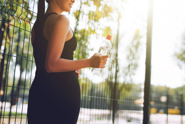 Siyah spor giysili genç bir kadın parkta esniyor ve spor yapıyor. Çekici, formda bir kızın bir şişe suyla dikilmiş görüntüsü. Sağlıklı yaşam tarzı kavramı. - Fotoğraf, Görsel