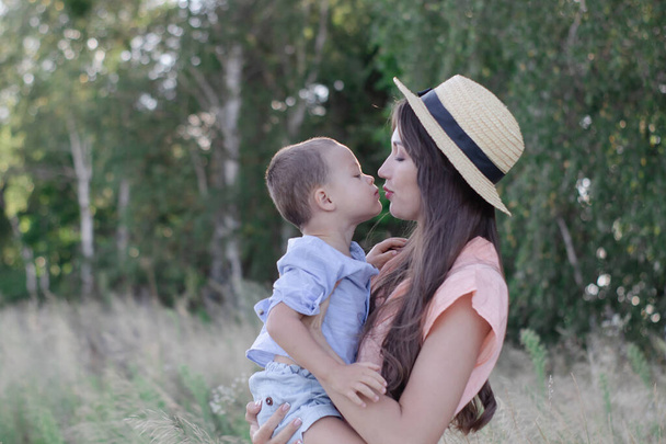 gelukkige familie - moeder en zoon knuffelen in het veld. mooie brunette in crème jurk en strohoed en kleine jongen in blauw kostuum. ouderschap, moederschap - Foto, afbeelding