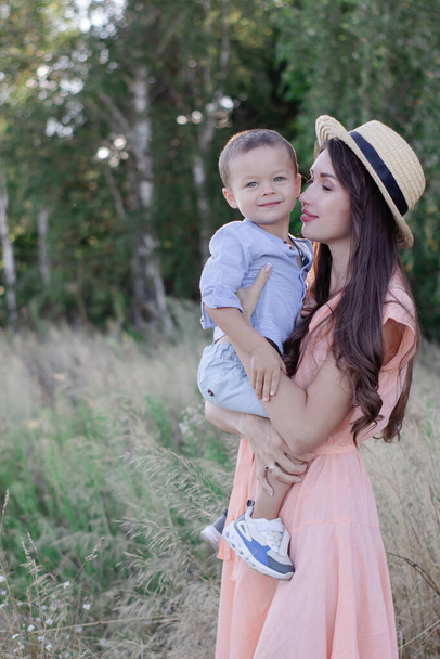 happy family - mother and son hug in the field. pretty brunette woman in cream dress and straw hat and little boy in blue costume. parenting, motherhood - Photo, Image
