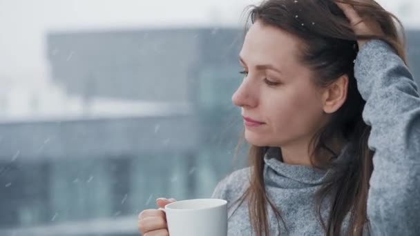 Femme reste sur le balcon pendant les chutes de neige avec une tasse de café chaud ou de thé. Elle regarde les flocons de neige et respire l'air frais glacé. - Séquence, vidéo