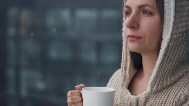 Mulher fica na varanda durante a queda de neve com xícara de café quente ou chá. Ela olha para os flocos de neve e respira no ar fresco gelado. - Filmagem, Vídeo