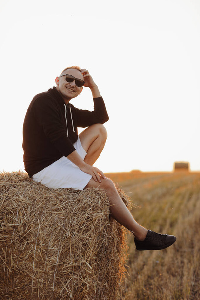 knappe jongeman in zonnebril, casual zwart sweatshirt en wit kort, in de zomer veld buiten, zittend op hooibaal, hooiberg op zonsondergang. - Foto, afbeelding