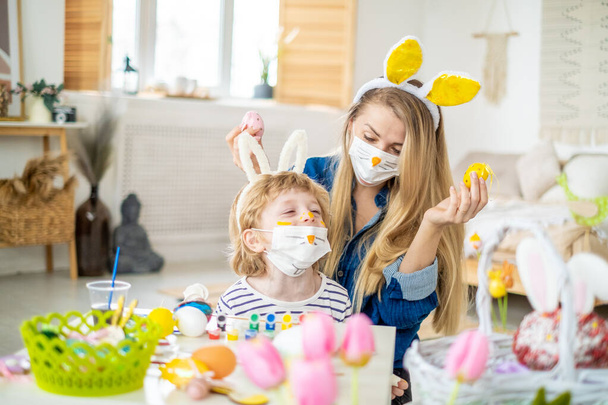 Happy cute mother and son wearing decorated bunny face mask and headbands with ears having fun getting ready for Easter holiday, family spend time at home during quarantine - Fotó, kép