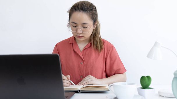 Aziatisch zakenvrouw in een helder t-shirt werkt thuis, Blijf thuis, Nieuw normaal, Covid-19 coronavirus, Afstanden op sociaal gebied, Ideeën voor werk bedenken, Informatie schrijven in een notitieboek. - Foto, afbeelding