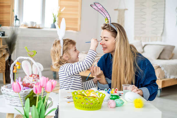 Красивый счастливый сын и мама в повязках с кроличьими ушами украшают яйца кисточкой и яркими красками, готовятся к празднованию Пасхи, веселятся дома - Фото, изображение