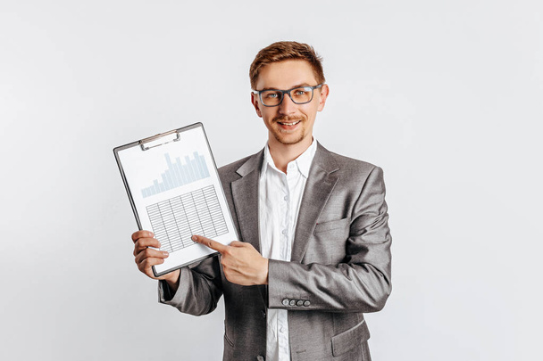 Young handsome brunet man in glasses in suit smiles and points at business with documents and graphs on white isolated background - Foto, Bild