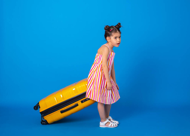 arabska dziewczynka w kolorowych ubraniach zmęczona noszeniem żółtej walizki na niebieskim tle. - Zdjęcie, obraz