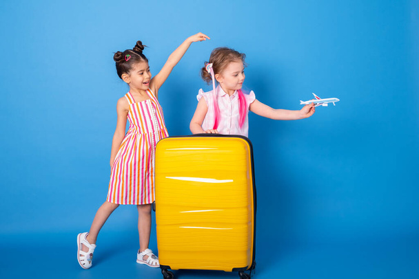 Happy excited little girls friends in colorful clothes with toy plane and yellow suitcase on blue background - Foto, imagen