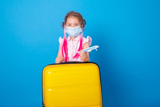 Muchacha divertida en máscara protectora con avión de juguete y maleta amarilla sobre fondo azul. - Foto, imagen