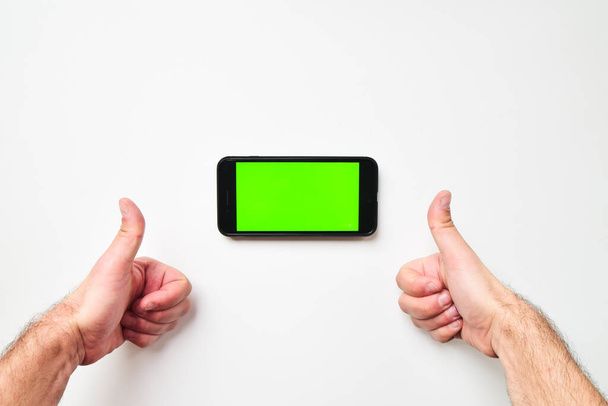 Mužské velké ruce a mobilní smartphone se zelenou obrazovkou v horizontální poloze na pozadí s dvěma palci nahoru prst. Vyhonit prázdné místo na obrazovce - Fotografie, Obrázek