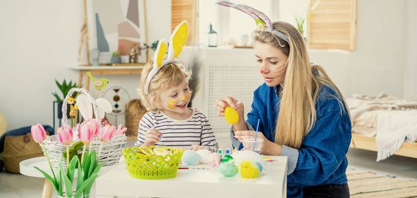 Beautiful happy son and mother in headbands with bunny ears decorate eggs with a brush and bright paints, prepare to celebrate Easter, have fun at home - 写真・画像
