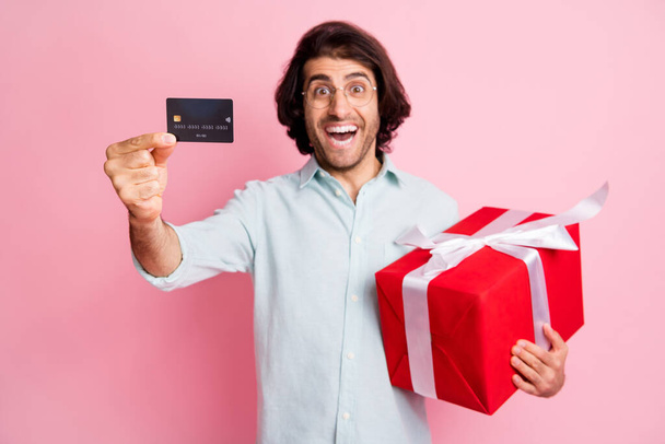 Portrait of young handsome happy overjoyed man male guy showing credit card hold red box gift wear glasses isolated on pink color background - Foto, Imagem