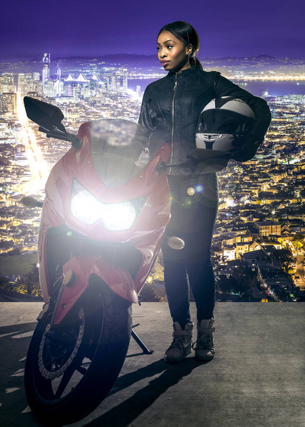 Sporty female motorcyclist or biker wearing a leather jacket and helmet on a road at night with a motorcycle above a scenic view of the San Francisco cityscape - Zdjęcie, obraz