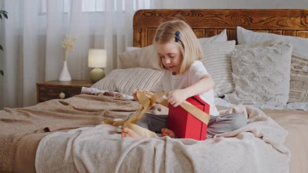 Nieuwsgierig klein meisje gelukkig kleuter blond dochter schattig baby kind zitten op gezellig bed met feestelijke rode geschenkdoos uitpakt aanwezig unties geel lint ziet er binnen, home vakantie verjaardagsfeest - Video