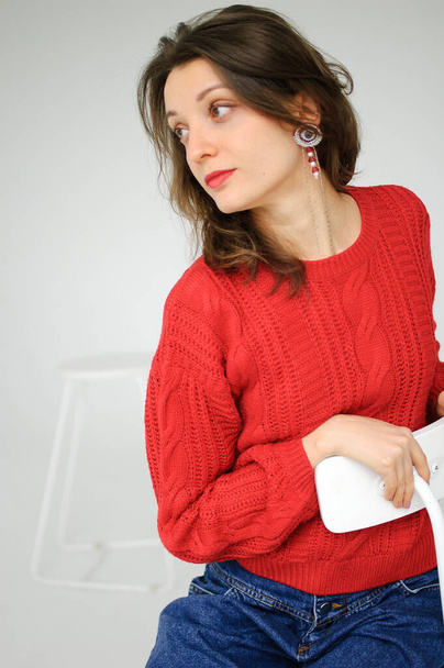 Stúdió portré fiatal nő piros kötött pulóver és fülbevaló ül egy széken fehér alapon - Fotó, kép