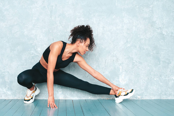 Portrait of fitness smiling black woman in sports clothing with afro curls hairstyle.She wearing sportswear.Young beautiful model stretching out before training.Female sitting in studio near gray wall - Foto, Imagen