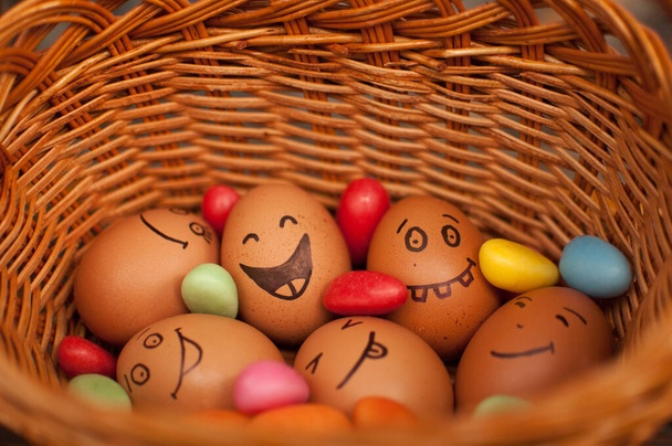 Stapel Eier mit handgezeichneten Gesichtern auf Strohkorb mit bunten Bonbons, Ostervorbereitung, Konzepte für Feiertagsstimmung - Foto, Bild