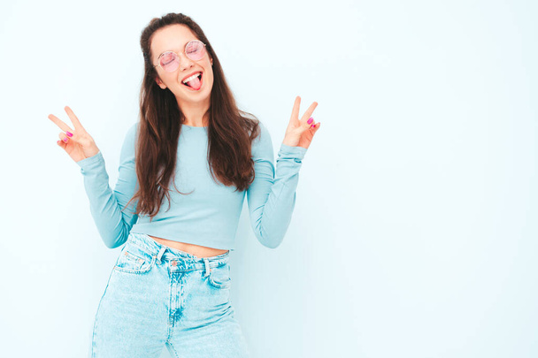 Young beautiful smiling female in trendy summer hipster clothes. Sexy carefree woman posing near light blue wall in studio. Positive model having fun indoors. Cheerful and happy.Shows peace sign - 写真・画像