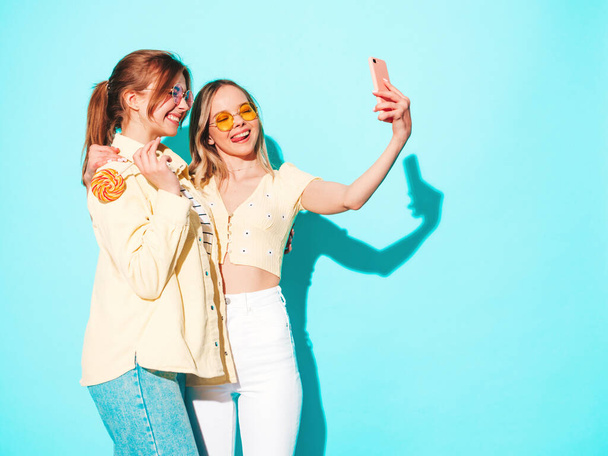 Two young beautiful blond smiling hipster female in trendy summer clothes. Sexy carefree women posing near blue wall in studio. Trendy and positive models having fun taking selfie - Fotó, kép