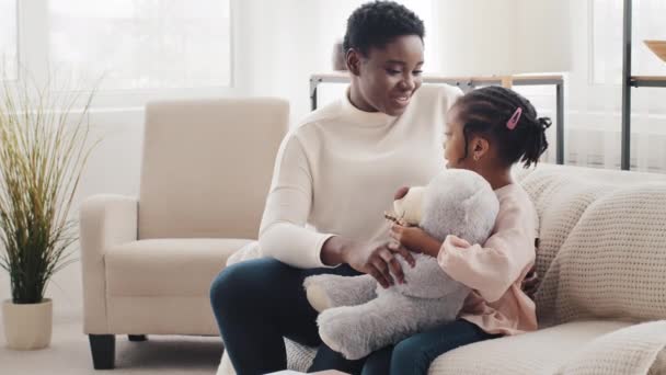 Счастливая африканская мать с афро маленькая дочь школьница сидит на диване дома говорить разговор, черная женщина мама няня общаться болтая с маленькой девочкой ребенок держит плюшевого мишку - Кадры, видео
