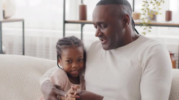 Little cute czarny dziewczyna córka wnuczka dziecko dziecko siedzi na kanapie z afro ojciec dojrzały tata etniczny afrykański dziadek mężczyzna przytulanie przytulanie czatowanie rozmowy oklaskiwanie klaskanie ręce śmiech - Materiał filmowy, wideo
