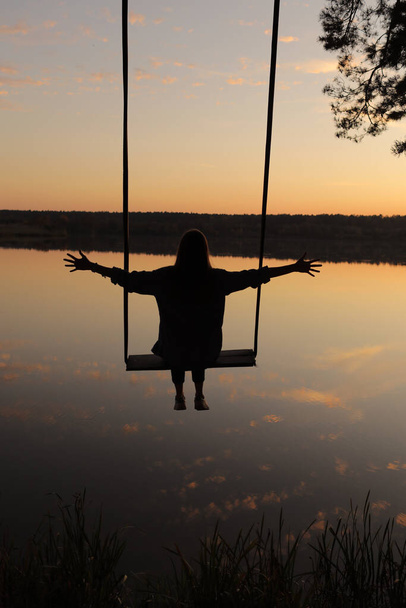 силует романтичної молодої жінки на гойдалці над озером на заході сонця. Молода дівчина мандрівник сидить на гойдалках в красивій природі, вид на озеро
. - Фото, зображення