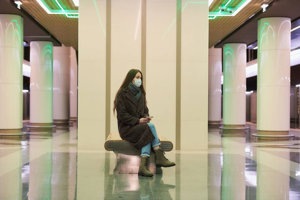 Una mujer solitaria con una máscara facial médica para evitar la propagación del coronavirus está sentada con un teléfono inteligente en la plataforma del metro. Una chica con una máscara quirúrgica mantiene una distancia social en el metro. - Foto, imagen