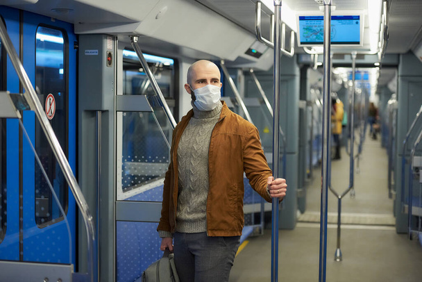 Egy szakállas férfi orvosi maszkban, hogy elkerülje a koronavírus terjedését, egy metrókocsiban ül és tartja a korlátot. Egy kopasz fickó sebészeti maszkban távolságot tart a vonaton.. - Fotó, kép