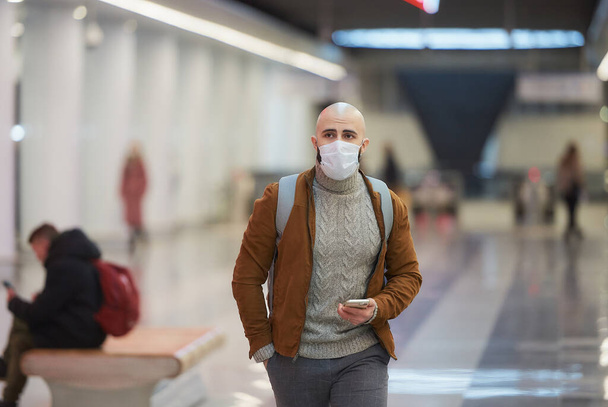 Ein Mann mit medizinischer Gesichtsmaske hält ein Smartphone in der Hand und läuft, während er in der Mitte der U-Bahn-Station auf einen Zug wartet. Ein Glatzkopf mit OP-Maske hält soziale Distanz. - Foto, Bild