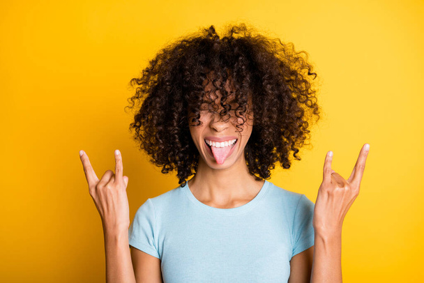 Foto van vrij grappige donkere huid krullende vrouw gekleed blauw t-shirt tonen hard rock teken tong geïsoleerde gele kleur achtergrond - Foto, afbeelding