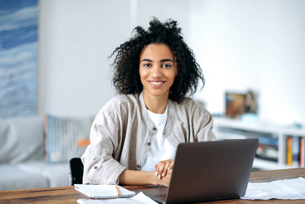 Schöne freundliche, selbstbewusste junge afrikanisch-amerikanische Frau in stylischer Freizeitkleidung, Freiberuflerin, Managerin oder Studentin, die am Laptop sitzt, zu Hause arbeitet oder studiert, in die Kamera schaut, lächelt - Foto, Bild