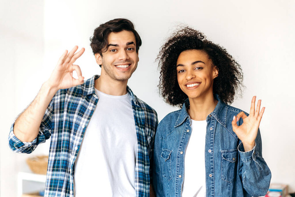 Portrait of young beautiful joyful mixed race couple, standing in casual wear at home, show with hands a sign okay, looking at the camera, smiling happily. A guy and a girl in love happy together - Photo, Image