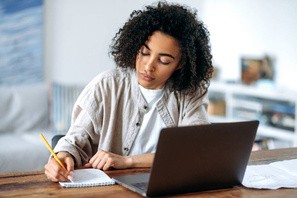 Focused confident smart african american curly haired girl in casual clothes, freelancer or student, working or studying remotely while sitting in living room at laptop and taking notes - Photo, Image