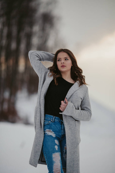 A young Caucasian female wearing a black shirt, jeans, and a jacket in a winter park covered in snow - Zdjęcie, obraz
