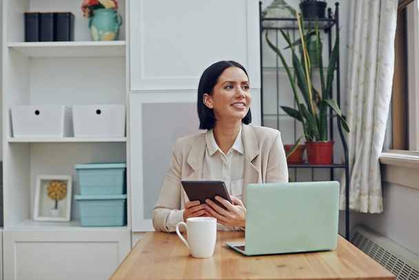 smiling businesswoman using laptop and looking at camera while sitting at table in office - Photo, Image