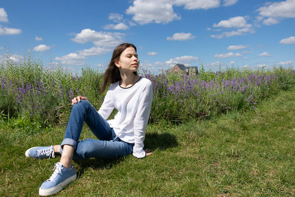Retrato de una joven con el pelo largo sentado en la hierba rodeada de flores de lavanda. La chica cerró los ojos y disfruta del aire fresco del país, la paz y la tranquilidad  - Foto, imagen
