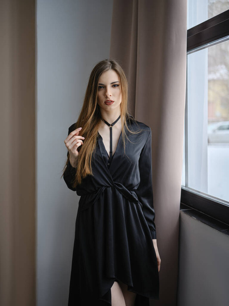 Young woman in black dress standing calm near the window - Фото, изображение