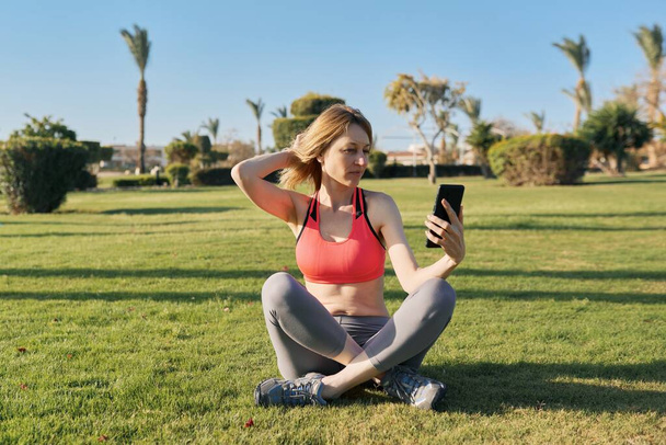 Mature woman doing morning exercises on green grass, with smartphone, video workout - Foto, afbeelding