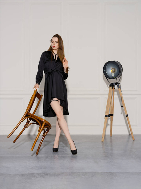 Playful young woman in short black dress leaning on the back of a chair with hands - Fotó, kép