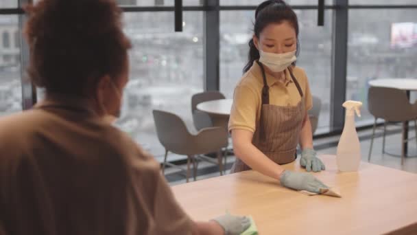 Sobre el hombro de un trabajador irreconocible del restaurante y una joven asiática con máscara médica y guantes, limpiando la mesa con trapos - Metraje, vídeo