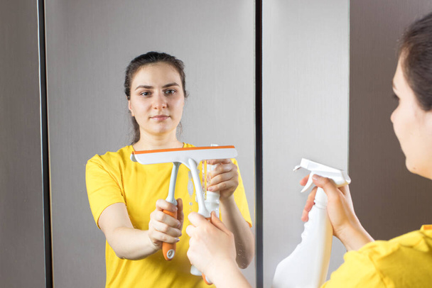 Η καθαρίστρια καθαρίζει τον καθρέφτη με ένα δοχείο νερού και καθαριστικό παραθύρων. Καθαρισμός σπιτιού. Υπηρεσία καθαρισμού - Φωτογραφία, εικόνα