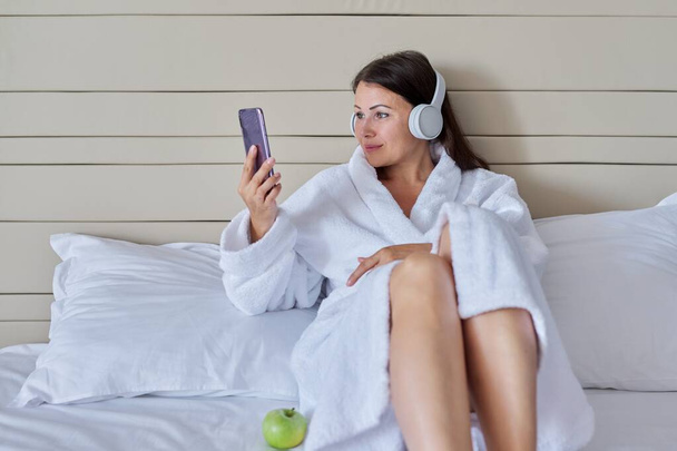 Happy mature woman in white bathrobe and headphones with smartphone sitting in bed - Фото, зображення