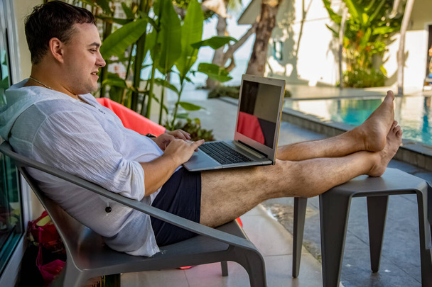 conceito de trabalho remoto. homem com laptop sentado à beira da piscina em um país tropical. - Foto, Imagem