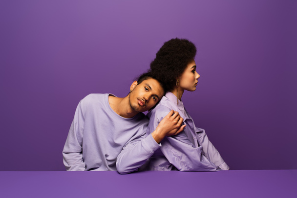 fashionable man leaning on young african american woman isolated on purple - 写真・画像