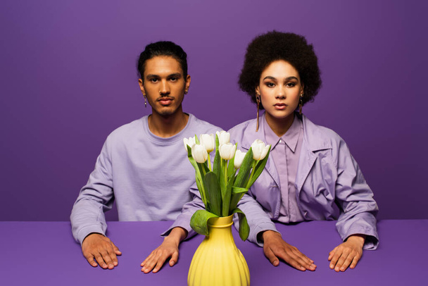Африканская американская пара сидит рядом с вазой с белыми тюльпанами изолированы на фиолетовый - Фото, изображение